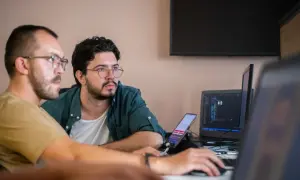 Dwóch programistów dyskutuje nad kodem. Jeden z nich wskazuje na ekran laptopa, gdzie widać kod w **JavaScript vs TypeScript**.