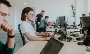 Młody programista w okularach skupiony na pracy przy laptopie. W tle inni pracownicy biura.