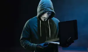 Haker w masce Guya Fawkesa, ubrany w bluzę z kapturem, pracuje przy laptopie. Symbolizuje zagrożenie w świecie bezpieczeństwa komputerowego.