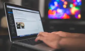 Ręce piszą na laptopie post "Jak promować moje nowe wpisy na blogu". Tło to rozmyty ekran telewizora z kolorowymi światłami, sugerujący relaks po pracy nad **cis benchmark**.