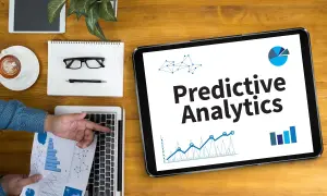Na tablecie widnieje napis "Predictive Analytics". Obok leżą laptop, notatnik z okularami i długopisem, filiżanka kawy i tablet.