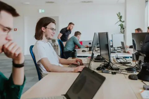 Młody programista w okularach skupiony na pracy przy laptopie. W tle inni pracownicy biura.