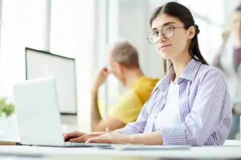 Młoda kobieta w okularach pracuje przy laptopie, szukając ofert pracy jako rpa developer.