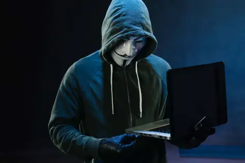 Haker w masce Guya Fawkesa, ubrany w bluzę z kapturem, pracuje przy laptopie. Symbolizuje zagrożenie w świecie bezpieczeństwa komputerowego.