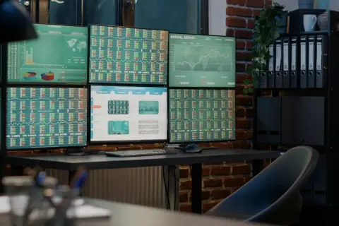 Biuro z wieloma monitorami wyświetlającymi dane finansowe i wykresy. Widać na nich wskaźniki zagrożeń (indicators of compromise) w postaci nietypowych wartości.