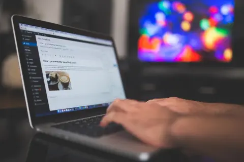 Ręce piszą na laptopie post "Jak promować moje nowe wpisy na blogu". Tło to rozmyty ekran telewizora z kolorowymi światłami, sugerujący relaks po pracy nad **cis benchmark**.