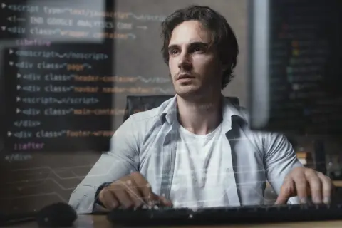 Młody mężczyzna, full stack developer, pracuje przy komputerze, na ekranie widać kod.