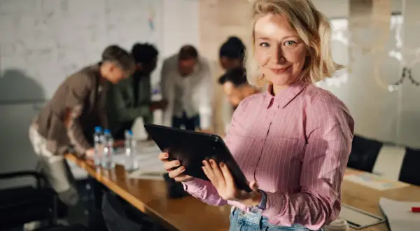 Kobieta z tabletem w dłoniach, uśmiecha się do kamery. W tle zespół dyskutuje przy stole. To inspiracja, jak zostać project managerem.