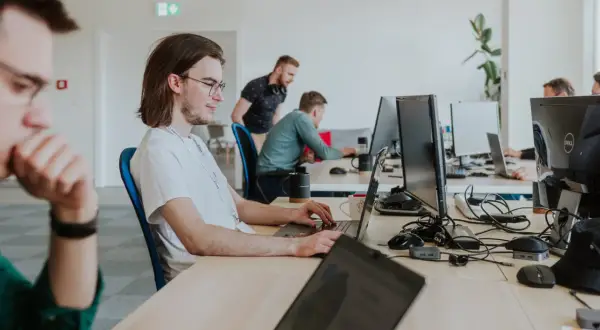 Młody programista w okularach skupiony na pracy przy laptopie. W tle inni pracownicy biura.