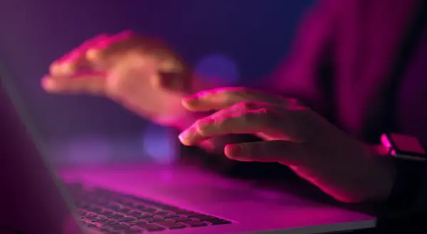 Dłonie nad klawiaturą laptopa w neonowym świetle. Czy to kodowanie, czy szukanie odpowiedzi na pytanie: uuid co to?