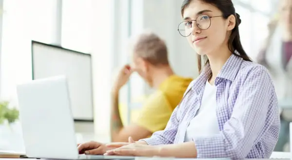 Młoda kobieta w okularach pracuje przy laptopie, szukając ofert pracy jako rpa developer.