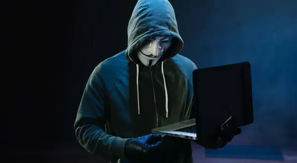 Haker w masce Guya Fawkesa, ubrany w bluzę z kapturem, pracuje przy laptopie. Symbolizuje zagrożenie w świecie bezpieczeństwa komputerowego.