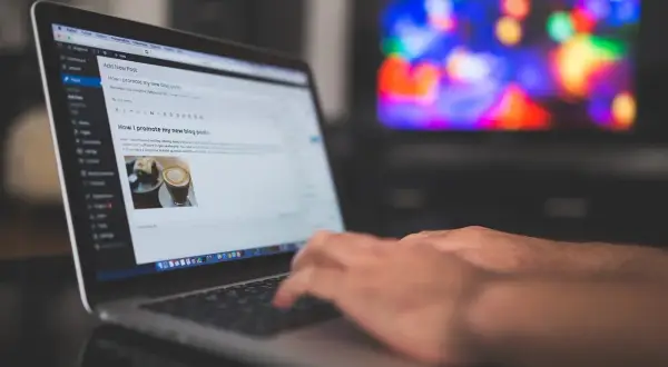 Ręce piszą na laptopie post "Jak promować moje nowe wpisy na blogu". Tło to rozmyty ekran telewizora z kolorowymi światłami, sugerujący relaks po pracy nad **cis benchmark**.