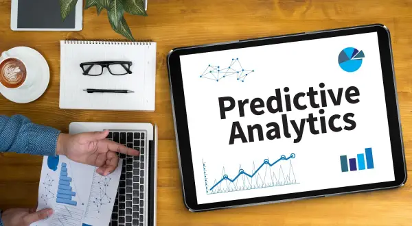 Na tablecie widnieje napis "Predictive Analytics". Obok leżą laptop, notatnik z okularami i długopisem, filiżanka kawy i tablet.