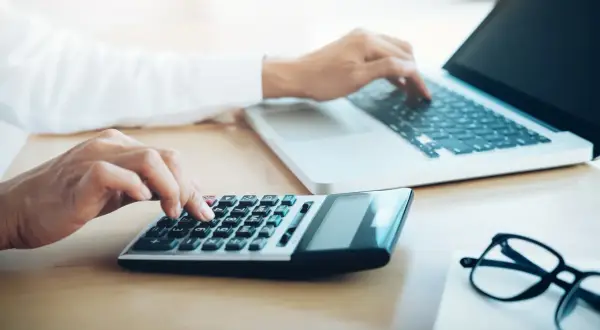 Ręce wpisują dane na kalkulatorze i laptopie, przygotowując się do zakupów e commerce.