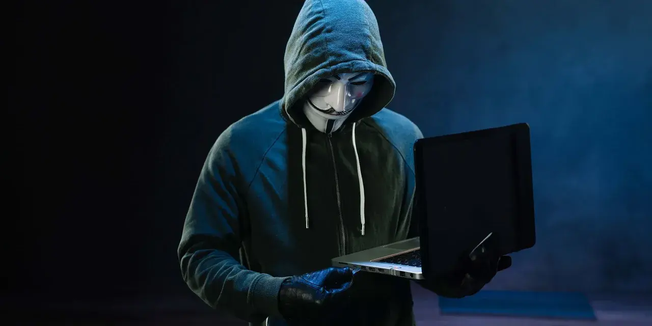 Haker w masce Guya Fawkesa, ubrany w bluzę z kapturem, pracuje przy laptopie. Symbolizuje zagrożenie w świecie bezpieczeństwa komputerowego.