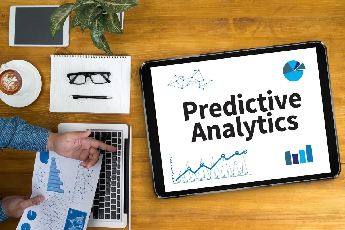 Na tablecie widnieje napis "Predictive Analytics". Obok leżą laptop, notatnik z okularami i długopisem, filiżanka kawy i tablet.