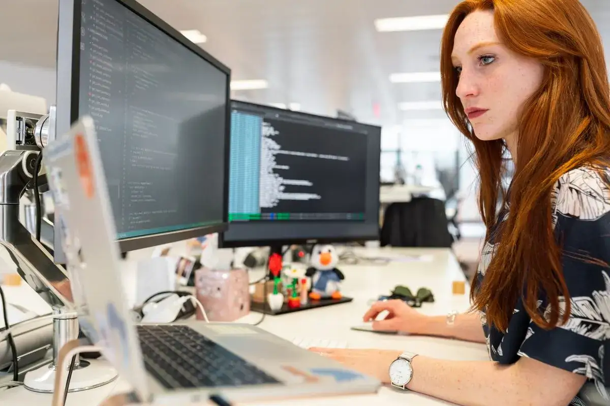 Młoda rudowłosa kobieta, junior IT project manager, pracuje przy komputerze, analizując kod na ekranie.