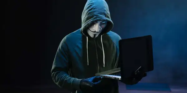 Haker w masce Guya Fawkesa, ubrany w bluzę z kapturem, pracuje przy laptopie. Symbolizuje zagrożenie w świecie bezpieczeństwa komputerowego.