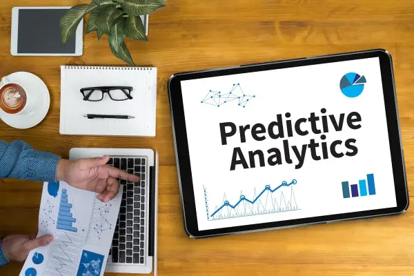 Na tablecie widnieje napis "Predictive Analytics". Obok leżą laptop, notatnik z okularami i długopisem, filiżanka kawy i tablet.